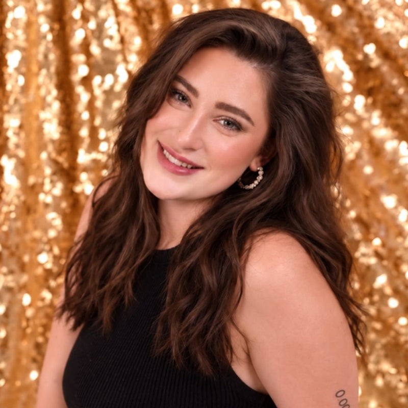 A woman with long wavy brown hair wearing a black ribbed sleeveless top, posing in front of a shimmering gold sequin backdrop.
